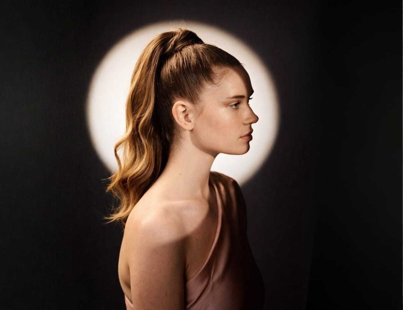 Profile of a woman with long hair in a ponytail against a spotlight backdrop.