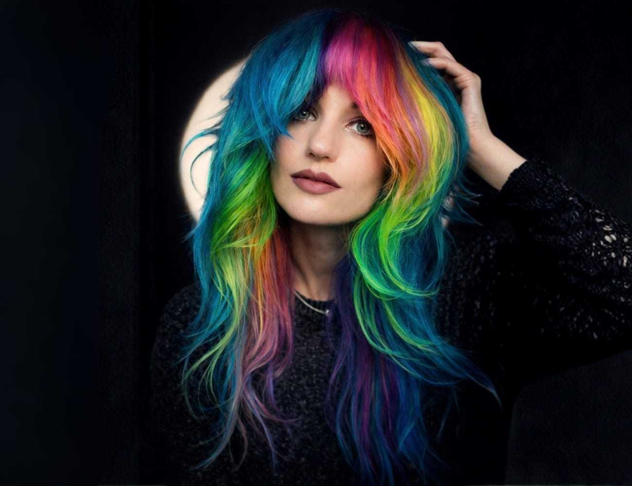 Young woman with vibrant rainbow-colored hair against a dark background.