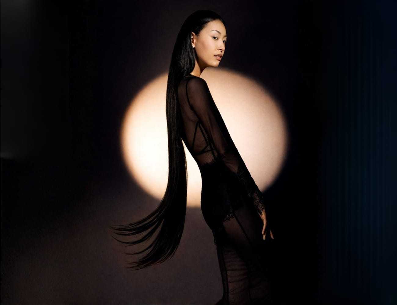 Woman in sheer black dress standing in spotlight with long, flowing hair.